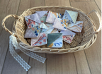 A wicker basket with handles sits on a light wood floor against a neutral wall, holding several small folded fabric pieces with colorful printed designs and white ribbon ties.