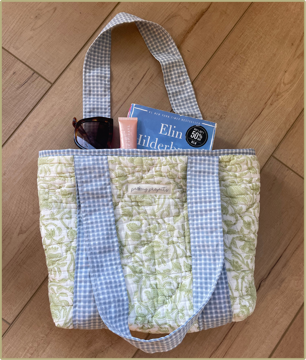 A quilted tote bag with a green floral pattern and blue-and-white gingham trim, shown on a wooden floor with sunglasses, a pink tube, and a book tucked inside.