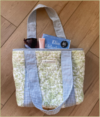 A quilted tote bag with a green floral pattern and blue-and-white gingham trim, shown on a wooden floor with sunglasses, a pink tube, and a book tucked inside.