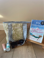 A quilted tote bag with a blue and white gingham pattern and a light green floral front panel, styled on a wooden floor next to a book on a small wooden stand, sunglasses, and two small toiletry items.