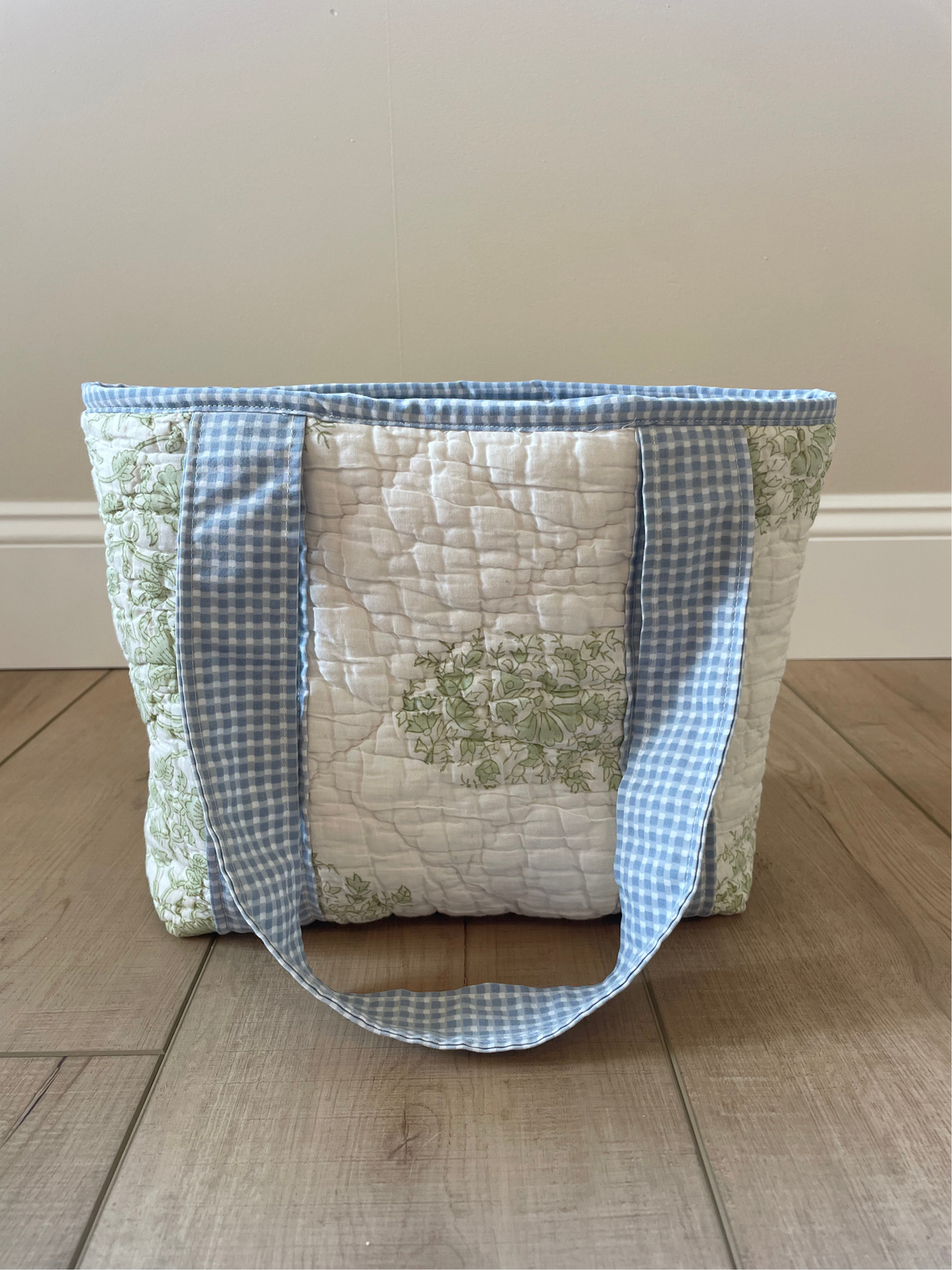 A handmade quilted tote bag with a central white quilted panel, light green floral print, and blue-and-white gingham fabric trim and handles, shown standing on a wooden floor against a beige wall.