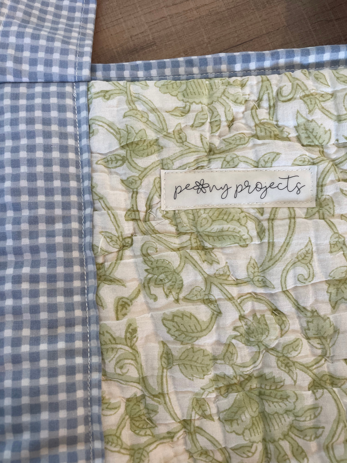 Close-up of a quilted fabric item showing a blue and white gingham border, a blue and white gingham top edge, and an off-white center panel with a green floral vine pattern. A sewn-on label reads 'peony projects'.