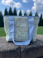 A quilted tote bag with a light green floral pattern and blue-and-white gingham straps, sitting on a stone wall outdoors with grass and trees in the background.