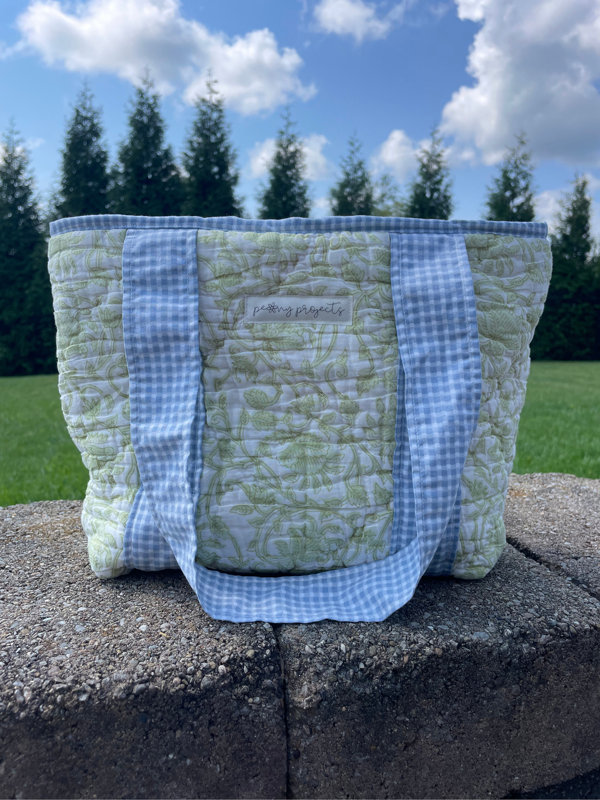 A quilted tote bag with a light green floral pattern and blue-and-white gingham straps, sitting on a stone wall outdoors with grass and trees in the background.