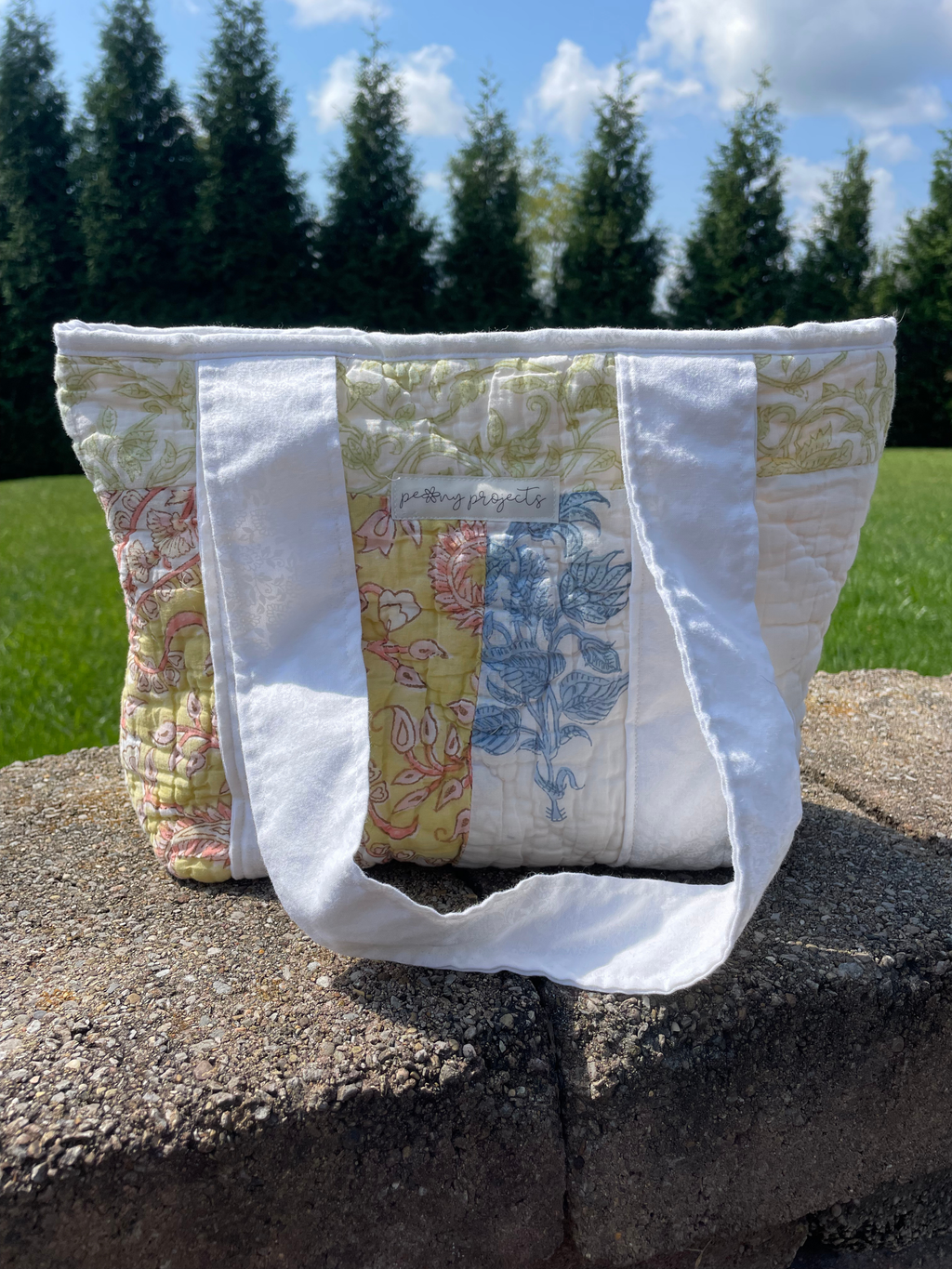 A handmade quilted tote bag with white fabric handles, featuring a patchwork design of green, yellow, and blue floral quilts, sitting on a stone wall outdoors with trees and grass in the background.