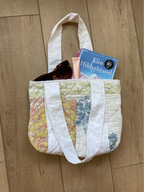 A handmade quilted tote bag with white canvas straps, filled with sunglasses, a pink tube, and a book, placed on a light wood floor.