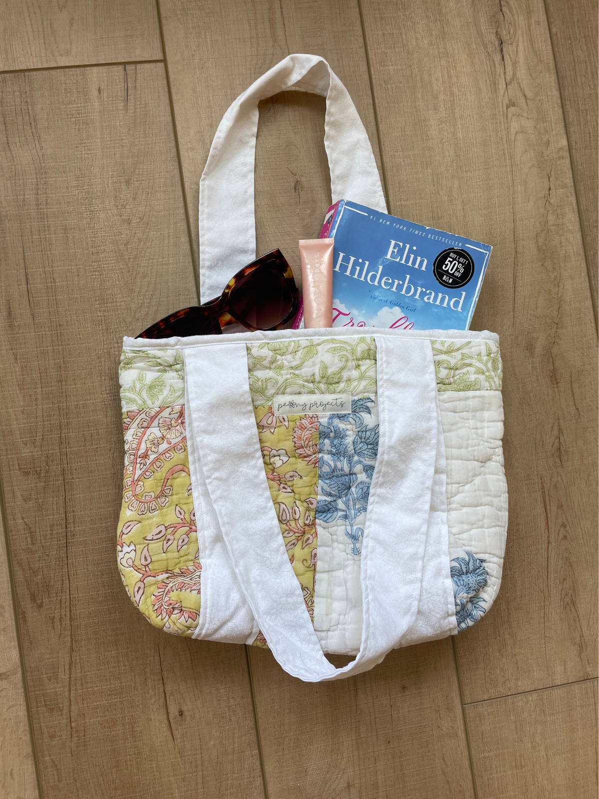 A handmade quilted tote bag with white canvas straps, filled with sunglasses, a pink tube, and a book, placed on a light wood floor.