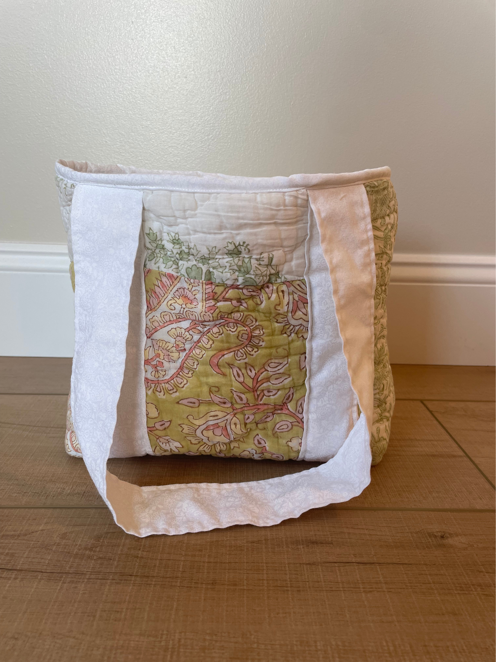 A handmade quilted fabric storage basket with a central floral pattern panel and white fabric handles, resting on a wooden floor against a beige wall.