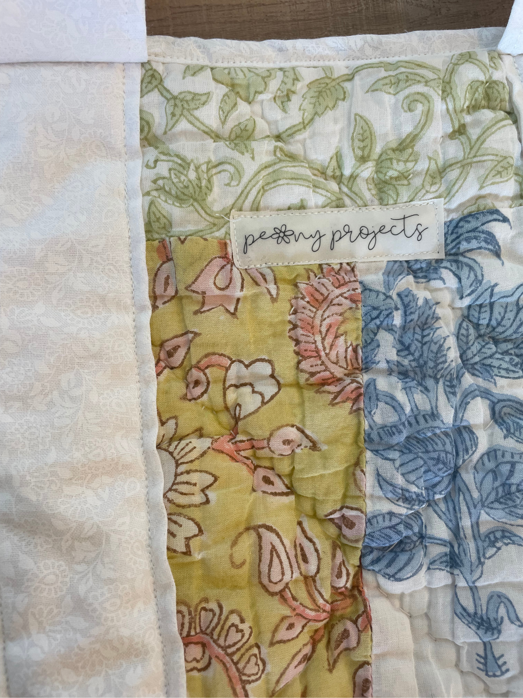 A close-up of a handmade quilted fabric piece with multiple patterned panels, including a central yellow panel with pink floral motifs, a blue botanical panel, and a cream panel with a visible 'peony projects' label.