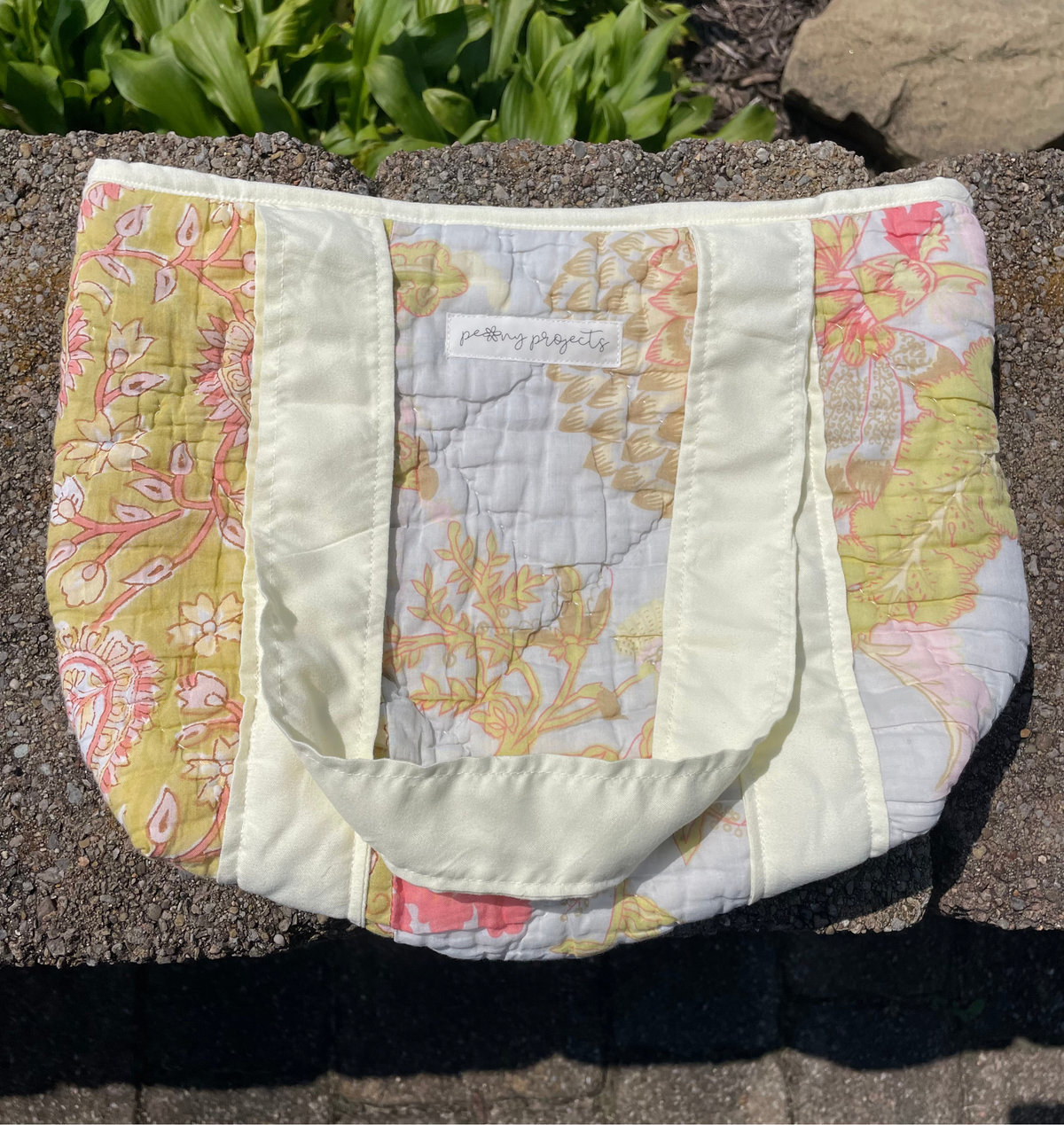 A handmade quilted fabric tote bag with a floral and paisley pattern, featuring cream-colored handles and trim, resting on a stone surface outdoors.