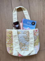 A quilted patchwork tote bag with cream handles and a floral patterned body, shown on a wood floor with books and sunglasses tucked inside.