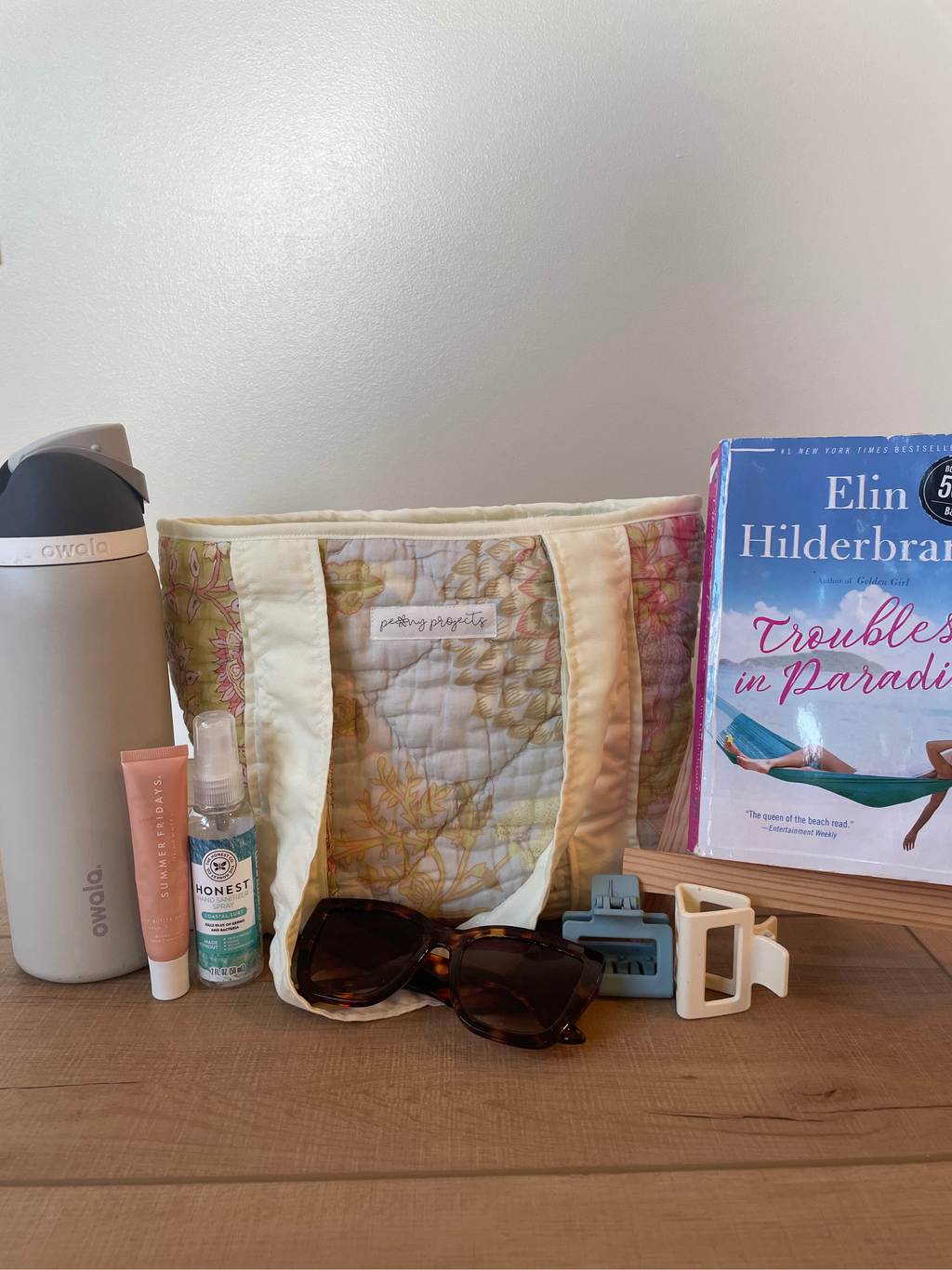 A quilted tote bag with a map print pattern sits on a wooden table surrounded by travel accessories, including a gray water bottle, a pink tube, a small spray bottle, tortoiseshell sunglasses, a blue card holder, and a book titled 'Trouble in Paradise' by Elin Hilderbrand.
