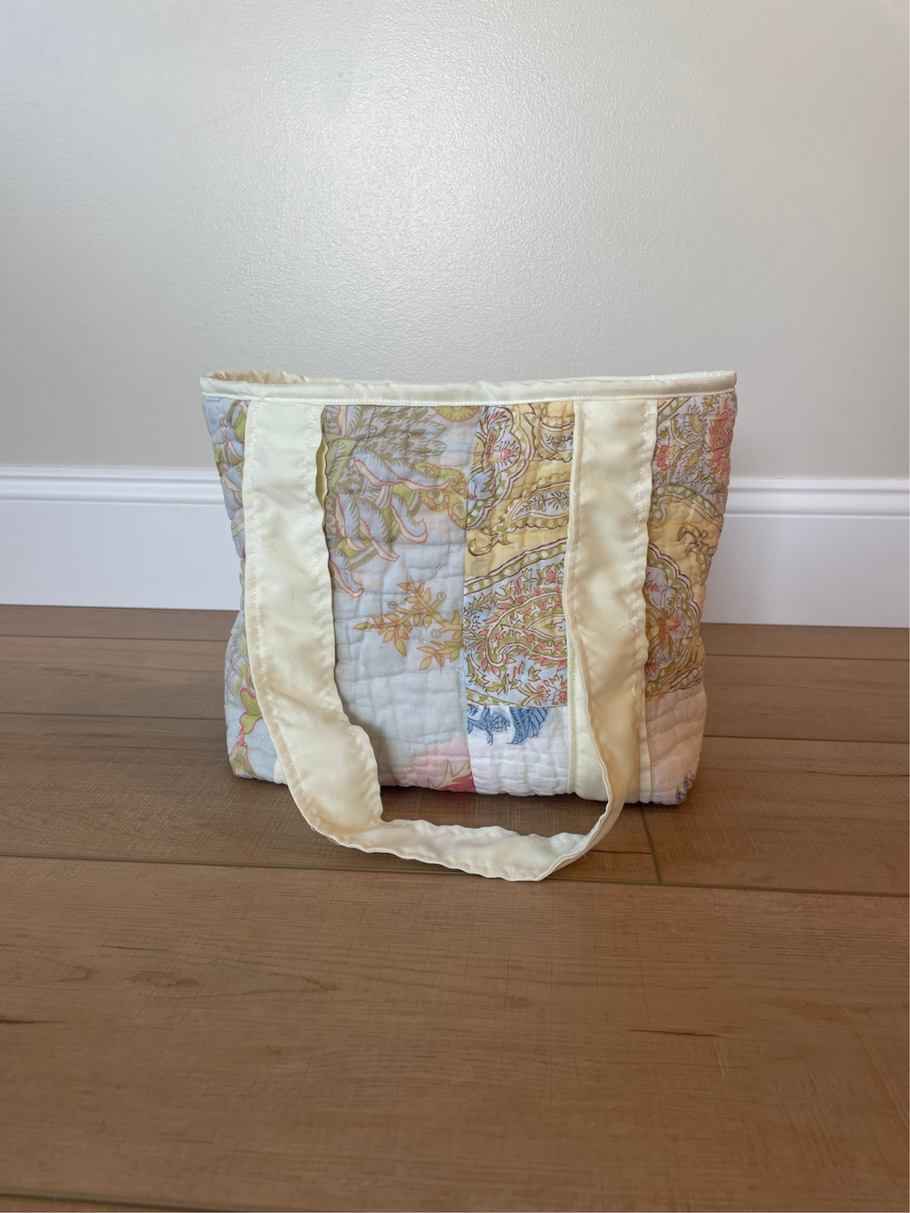 A quilted tote bag with a vintage-style map print in soft pastel colors and cream-colored satin straps, standing on a wooden floor against a beige wall.