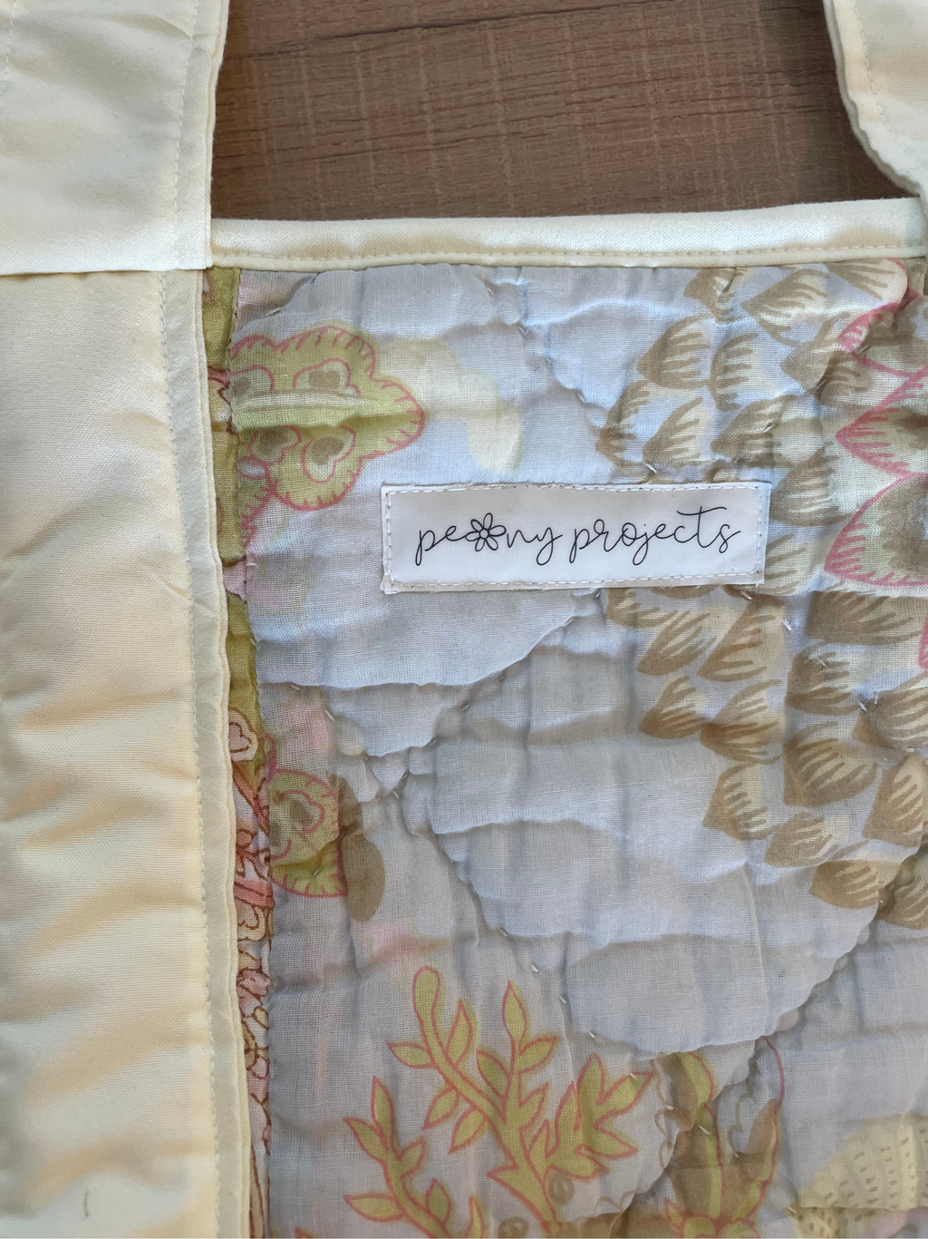 Close-up of a quilted fabric item with a cream satin border and a patterned center panel in soft beige, pink, and white tones. A sewn label reads 'petty projects', showing the brand.