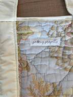 Close-up of a quilted fabric item with a cream satin border and a patterned center panel in soft beige, pink, and white tones. A sewn label reads 'petty projects', showing the brand.