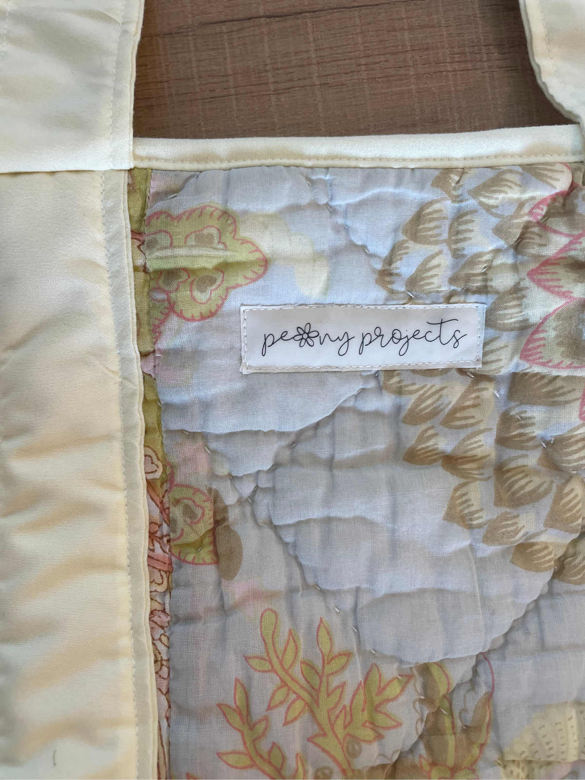 Close-up of a quilted fabric item with a cream satin border and a patterned center panel in soft beige, pink, and white tones. A sewn label reads 'petty projects', showing the brand.