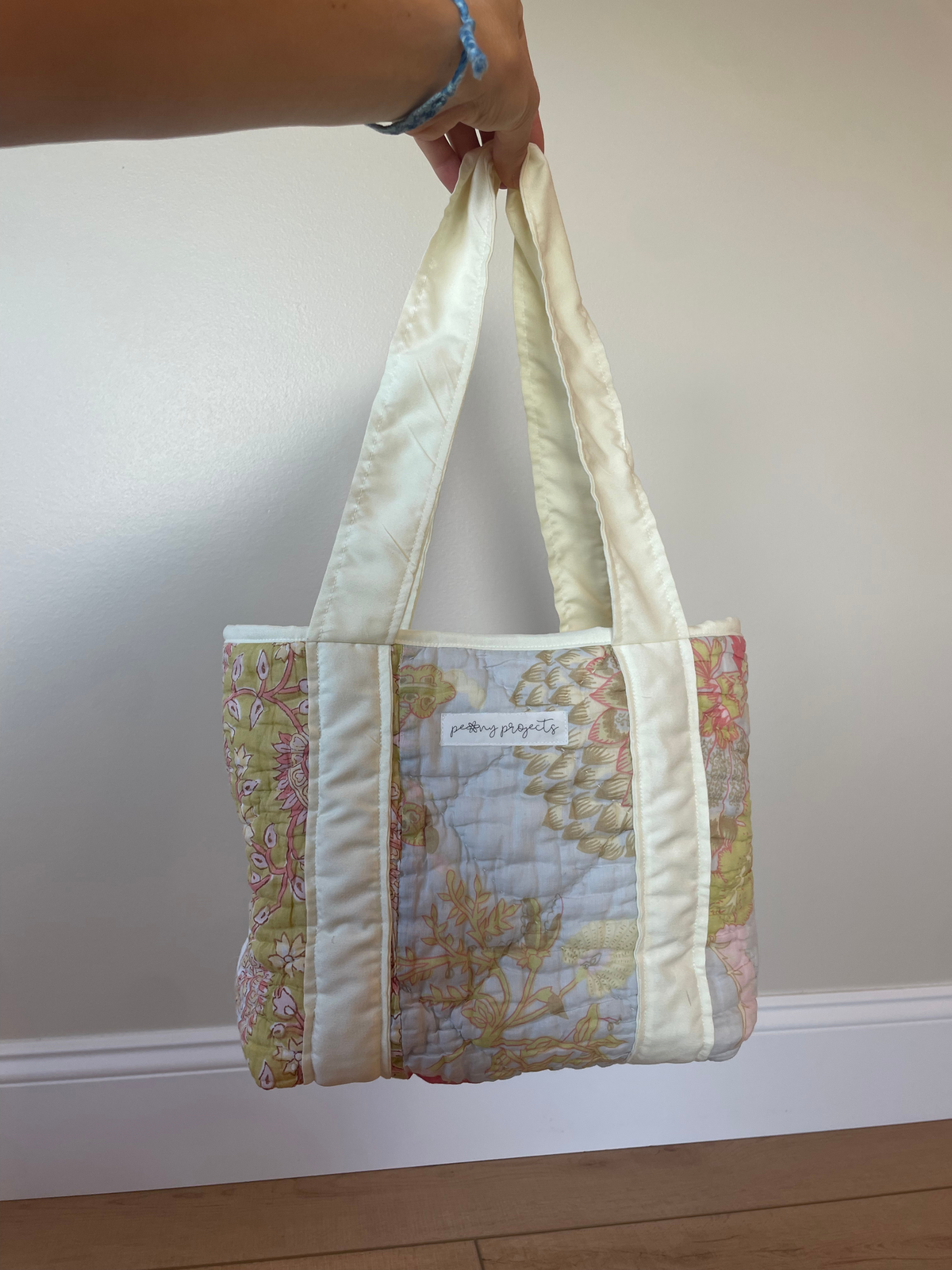 A handmade quilted tote bag with floral patterned panels and cream-colored handles, held by a person's hand against a plain indoor wall.