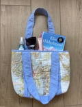 A quilted tote bag with a light blue fabric trim and a vintage map print body, shown filled with various items on a wooden floor. The bag features a map pattern with light blue borders and handles.