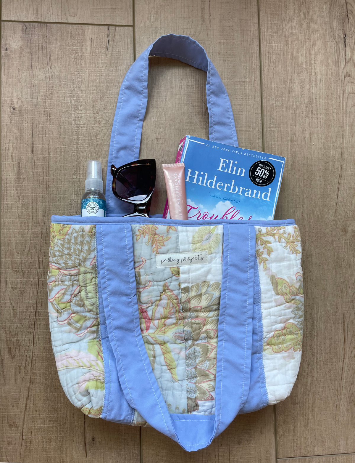 A quilted tote bag with a light blue fabric trim and a vintage map print body, shown filled with various items on a wooden floor. The bag features a map pattern with light blue borders and handles.