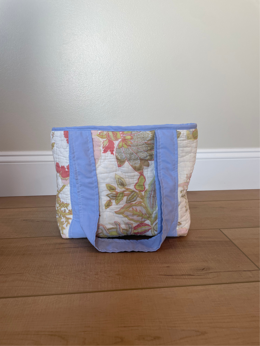 A handmade quilted tote bag with a floral pattern and light blue fabric trim, standing on a wooden floor against a plain wall.