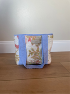 A handmade quilted tote bag with a floral pattern and light blue fabric trim, standing on a wooden floor against a plain wall.