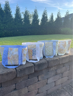 Four quilted fabric tote bags with blue handles and blue trim, lined up on a stone wall outdoors with a green lawn and trees in the background.