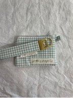 A small light green and white gingham check pouch or case with a gold-tone metal ring and strap, photographed on a wrinkled white fabric background.