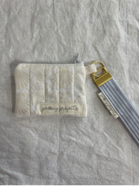 A small quilted cream zip pouch with a white-and-gray striped wrist strap and gold-tone hardware, laid on a wrinkled light fabric background.