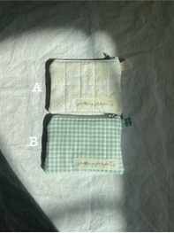 Two small zippered pouches or cases laid flat on a wrinkled white fabric surface under direct sunlight. The top pouch is off-white with a small stitched label, and the bottom pouch has a light green and white gingham pattern with a matching label.