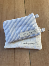 Two small fabric pouches or organizers stacked on a wood surface. The top pouch is light blue and white striped, and the bottom pouch is off-white.