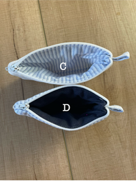 Two fabric pouches or small bags laid flat on a wood surface, labeled C and D. The top pouch has a white and gray striped fabric exterior with a white zipper and pull, and the bottom pouch has a dark navy interior.