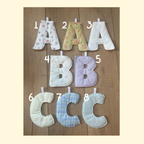 Eight soft fabric letter pillows shaped like the letters A through C, shown in multiple pastel patterns and colors on a wooden surface. The letters are numbered 1 through 8 with white digital text overlays.