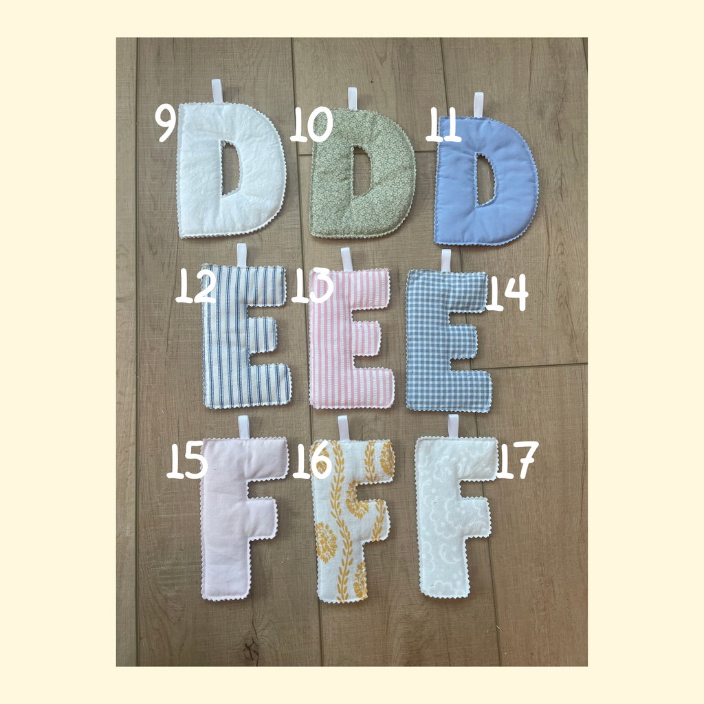 Seven handmade fabric letters D through F with numbers 9 through 17 printed in white near each letter, arranged on a wooden surface. The letters feature different patterns: white, sage green, light blue, blue and white stripes, pink and white stripes, light blue gingham, and white with a yellow floral print.
