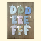 Seven handmade fabric letters D through F with numbers 9 through 17 printed in white near each letter, arranged on a wooden surface. The letters feature different patterns: white, sage green, light blue, blue and white stripes, pink and white stripes, light blue gingham, and white with a yellow floral print.