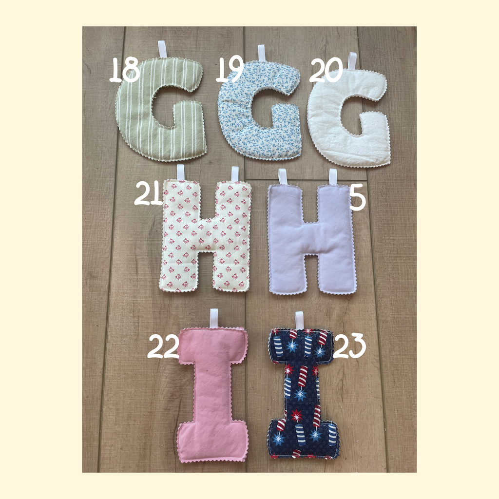 Six fabric letter pillows arranged on a wood surface, each labeled with a number from 18 to 23. The letters include G, H, and I in multiple pastel and patterned fabrics, including stripes, florals, solid lavender, and a navy patriotic print.