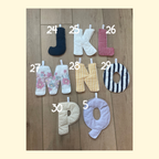 Nine fabric letter letter pads from J to Q arranged on a wooden surface, each numbered with a white handwritten number. The letters feature various patterns including solid navy, white, red gingham, floral, yellow gingham, black and white stripes, cream bouclé, and light lavender.