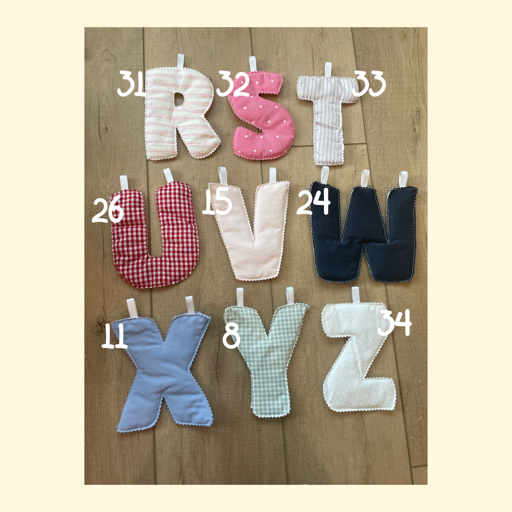 Nine quilted fabric letters A through Z arranged in a 3x3 grid on a wooden surface, each with a white clip at the top. The letters are in different colors and patterns, including pink polka dots, red gingham, light blue, mint green gingham, and white.
