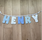 A fabric letter banner spelling 'HENRY' hanging on a white string against a light brown wooden floor.