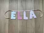 A handmade fabric letter banner spelling 'BELIEVE' in vertical orientation, hanging from a pink cord against a light brown wooden floor.