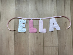 A handmade fabric letter banner spelling 'BELIEVE' in vertical orientation, hanging from a pink cord against a light brown wooden floor.