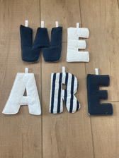 Fabric letters spelling 'WE ARE' arranged on a light wood floor, with navy blue, white, and black-and-white striped letters.