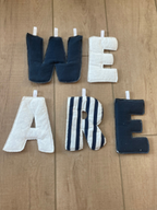 Fabric letters spelling 'WE ARE' arranged on a light wood floor, with navy blue, white, and black-and-white striped letters.