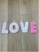 Four fabric letters spelling LOVE arranged on a light wood floor, with the first two letters in pale pink and the last two in bright pink, each clipped to the floor.