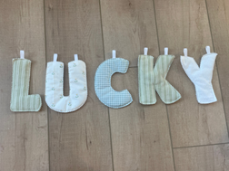 A set of fabric letter cutouts spelling 'LUCKY' arranged vertically on a light wood floor. The letters are made from soft fabrics in pale green, cream, and white gingham patterns, with white plastic tabs at the back.