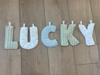A set of fabric letter cutouts spelling 'LUCKY' arranged vertically on a light wood floor. The letters are made from soft fabrics in pale green, cream, and white gingham patterns, with white plastic tabs at the back.