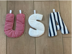 Three fabric letter pillows spelling 'USA' arranged vertically on a light wood floor. The top letter is red and white gingham, the middle is solid white, and the bottom is navy and white striped.