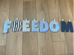 A decorative banner spelling out the word 'FREEDOM' in large letters, made from blue and white striped and solid navy fabrics, laid out on a light brown wooden floor.