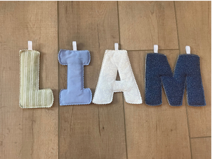 A fabric letter banner spelling 'I AM' laid out on a wooden floor. The letters are made of soft woven fabric in cream, light blue, white, and dark blue denim-like material, each attached to a white plastic hook.