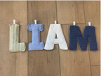 A fabric letter banner spelling 'I AM' laid out on a wooden floor. The letters are made of soft woven fabric in cream, light blue, white, and dark blue denim-like material, each attached to a white plastic hook.