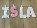 Three handmade quilted fabric letter cushions spelling 'SUS' arranged vertically on a light wood floor. The top cushion is white with a small pink floral pattern, the middle cushion is solid light pink, and the bottom cushion is white with thin pink stripes.